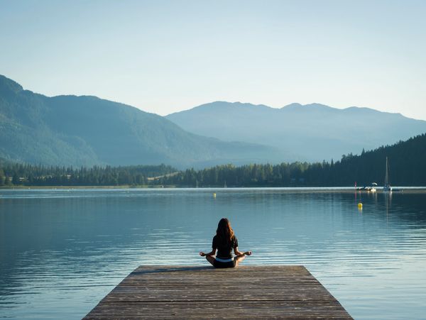 A woman meditating on a dock overlooking a calm lake and mountains.