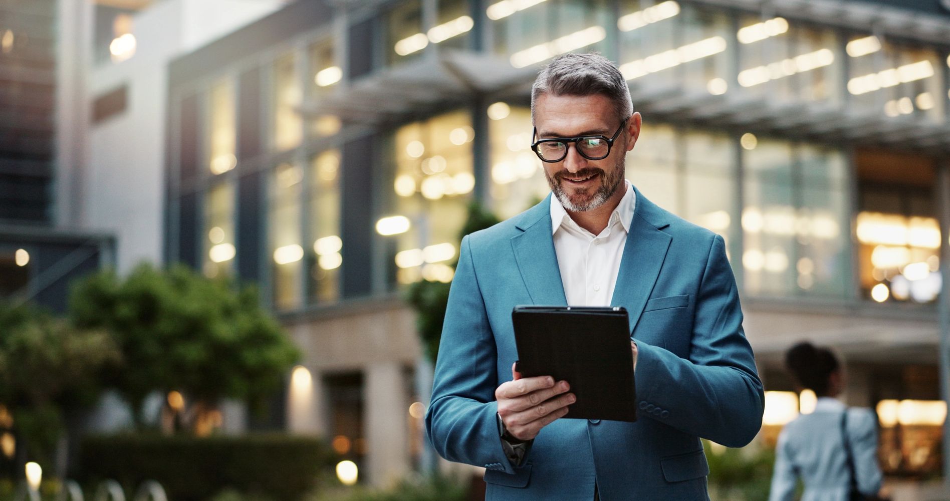 Smiling businessman using a tablet outside a modern office building.