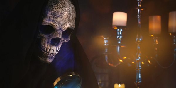 A figure in a skull mask and dark cloak under candlelight.