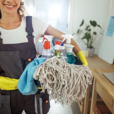 Smiling cleaner holding cleaning supplies and mop in a bright room.