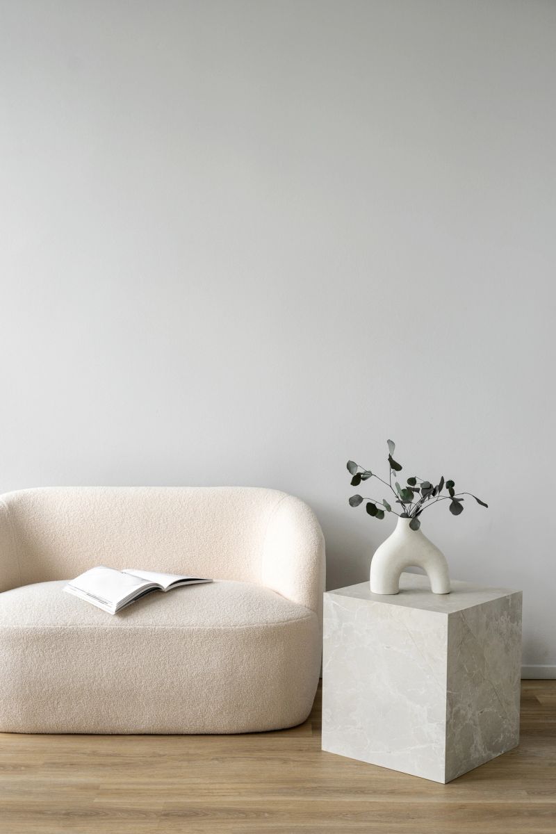 Vertical shot of white couch and side table are featured in a minimal interior setting. Vase with greenery close to sofa with open book in living room with copy space wall on backgrounds. Home design
