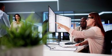 Woman points at computer screen while discussing work with colleague.