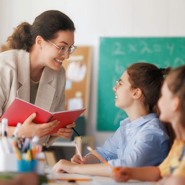 Teacher engaging with students in a classroom, holding a red notebook.