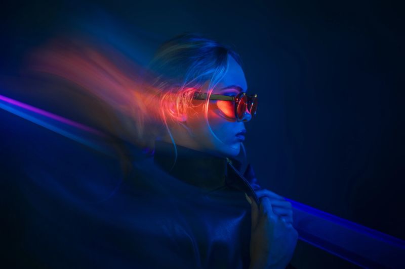 Profile of a woman wearing sunglasses, illuminated by vibrant neon lights, creating a futuristic, cyberpunk-inspired atmosphere with motion effects.