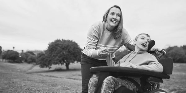 Woman smiling and pushing boy in wheelchair outdoors.