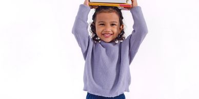 Smiling child balancing books on head, wearing a purple sweater and blue pants.