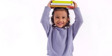 Smiling child balancing books on head, wearing a purple sweater and blue pants.