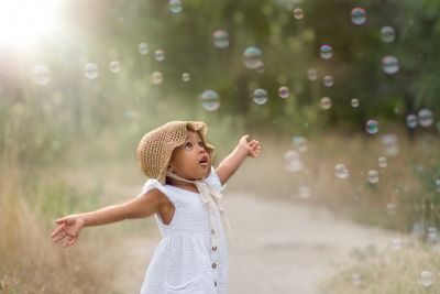 A toddler in a straw hat reaching out to bubbles outdoors.