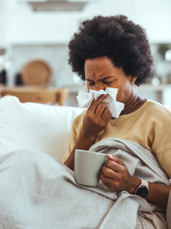 A woman blowing her nose while holding a mug, wrapped in a blanket.