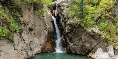 A small waterfall cascades into a clear pool surrounded by rocky cliffs and green foliage.