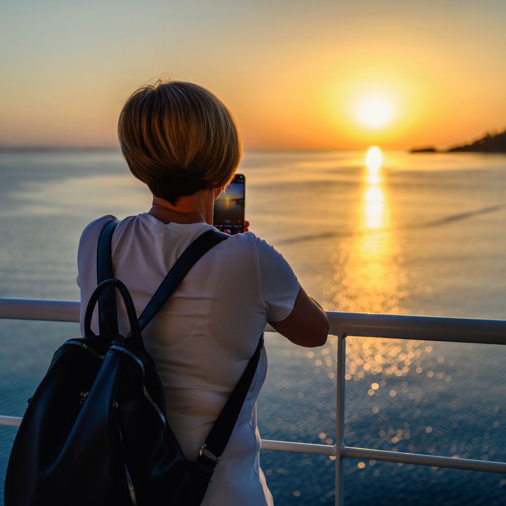 Woman with backpack capturing a sunset over calm water with her phone.