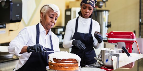 Two women baking and decorating a layered cake in a kitchen.