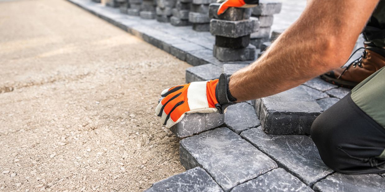 Worker laying gray paving stones on sandy ground wearing orange gloves.