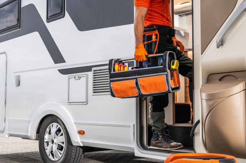 A technician wearing an orange shirt and work pants is entering a motorhome while carrying a toolbox. The toolbox is full of tools, including screwdrivers and other items. The motorhome is parked outdoors on pavement.