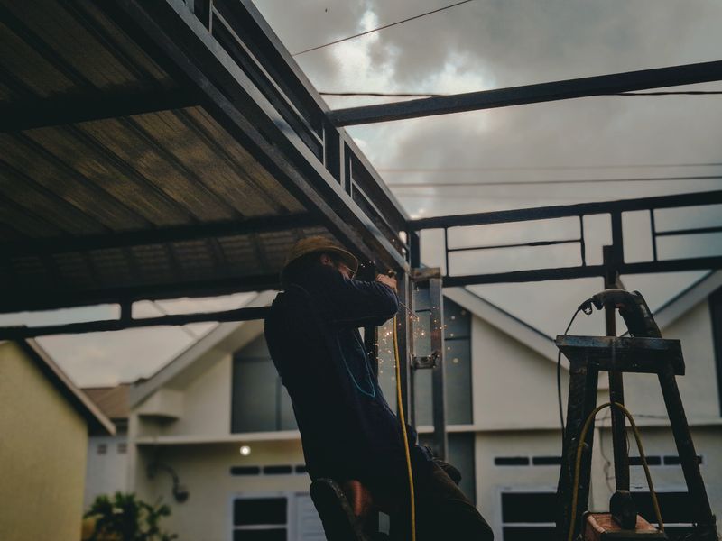 This photo captures an individual engaged in welding work on a metal structure, The serene backdrop of a building and a clear sky, likely at dawn or dusk, contrasts beautifully with the intense manual labor and skilled trade being showcased