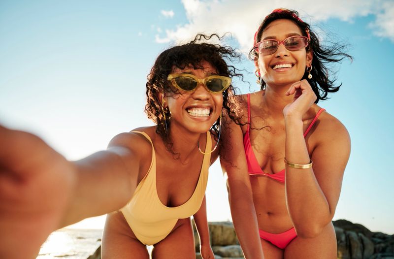 Portrait of two laughing young female friends in swimwear taking a selfie together during a sunny day at the beach in summer
