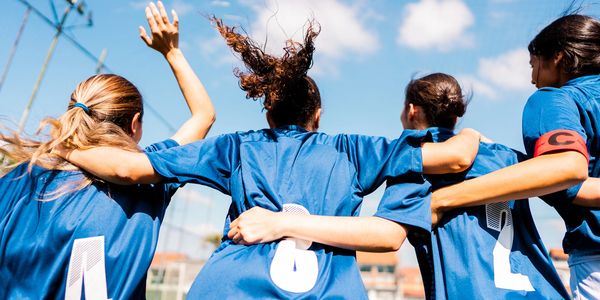 Girls in blue sports jerseys cheer with arms around each other under a sunny sky.