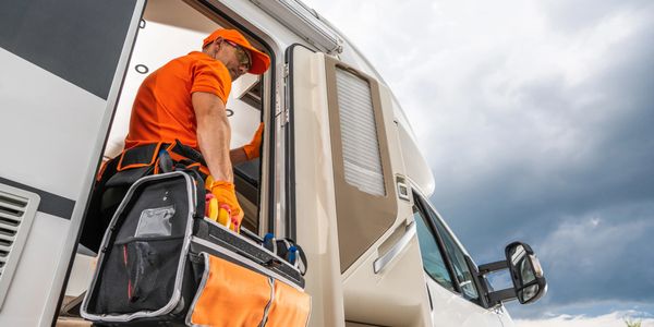 Technician in orange uniform carrying a toolbox into an RV under cloudy skies.