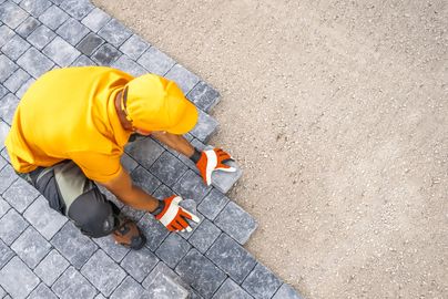 Worker laying gray paving stones on the ground wearing orange gloves and cap.