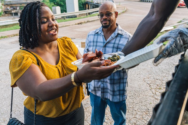 The Compostable box has Smoked Pork Sausage, BBQ Chicken, Baked Beans, and Dirty Rice.

Black Entrepreneur Small Business Food Truck Owner Man Setting Up, Hosting, and Serving Customers in Downtown Houston, Texas, USA. Part of a Series, Photographed in Houston, Texas.