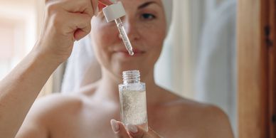 A woman applies a skincare serum with a dropper, wearing a towel on her head.