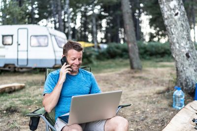 Man camping while talking on phone and looking at laptop computer. 