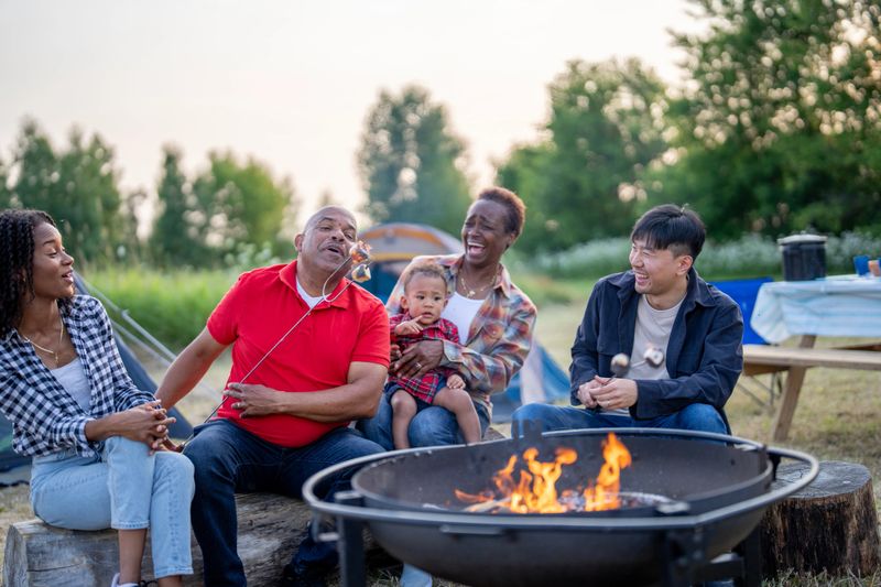 A small family of five sit around a fire during a family camping trip as they roast marshmallows together.  They are each dressed casually and are laughing as one family member accidentally catches his marshmallow on fire.