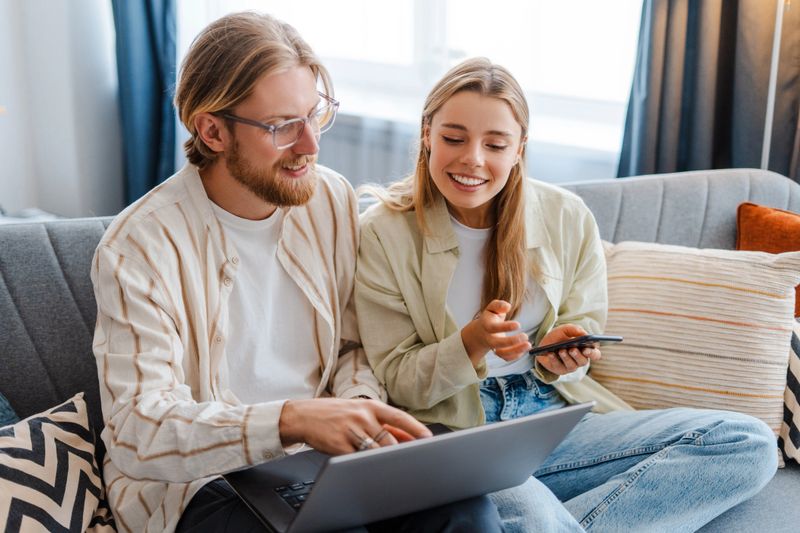 Young couple is sitting on a sofa at home, browsing on a laptop and checking prices on a smartphone. They are smiling and enjoying their time together. Online technology concept