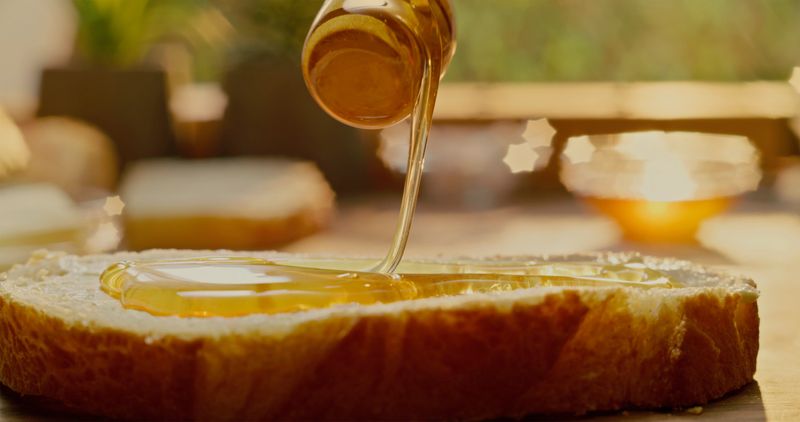 A close-up shot capturing honey being drizzled onto a thick slice of bread, with warm, golden sunlight softly illuminating the scene.