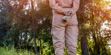 Person in beige outfit holding gloves behind back in a sunlit forest.