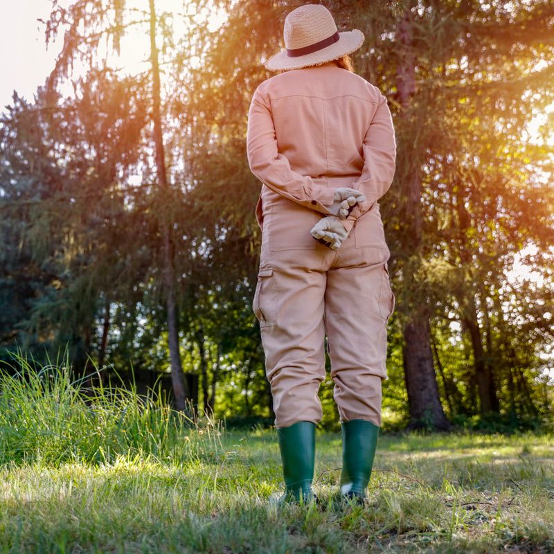 Woman gardener in straw hat and brown garden suit  in the garden in warm autumn sunny day. Lush vegetation and grass and trees background. Autumn gardening.