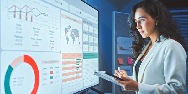 A woman analyzing data on a large digital screen while taking notes on a tablet.