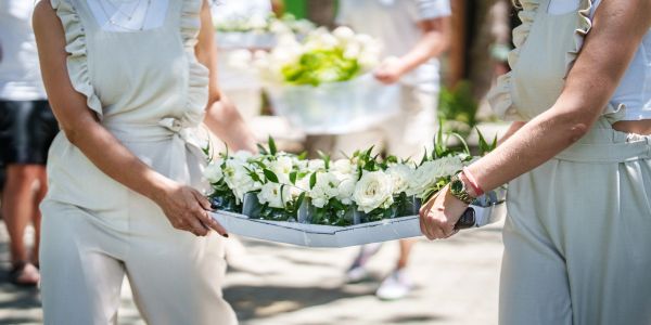 Two people in white carrying a tray with white roses and greenery.