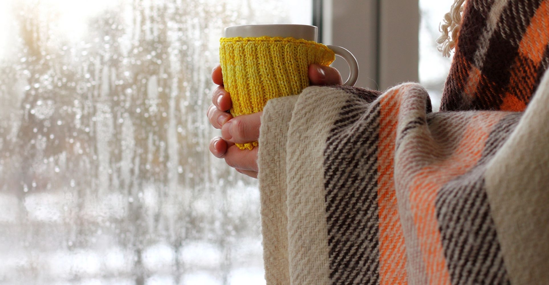 Person wrapped in a cozy blanket holding a cup by a rainy window.