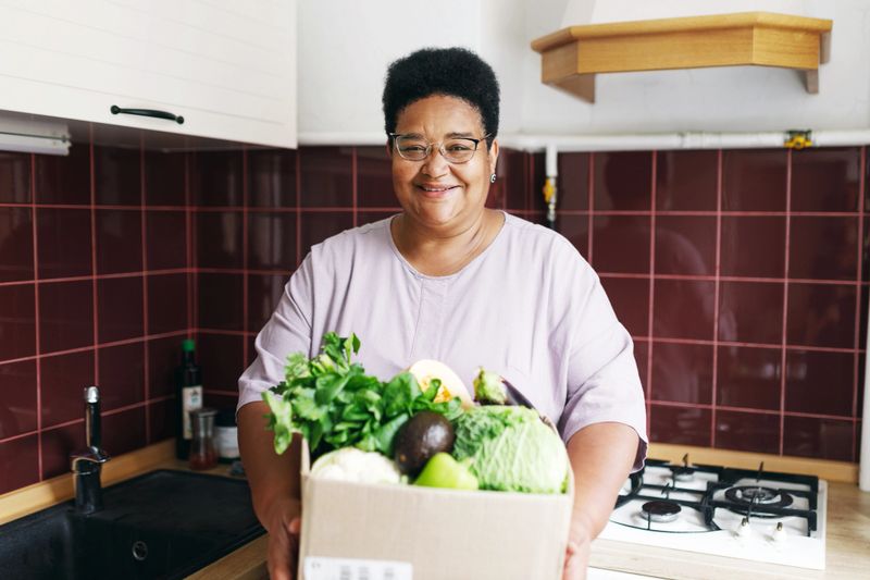Happy smiling contented overweight african american woman of 60s holding big card box with fresh vegetables standing at kitchen against counter, after receiving her food delivery service order