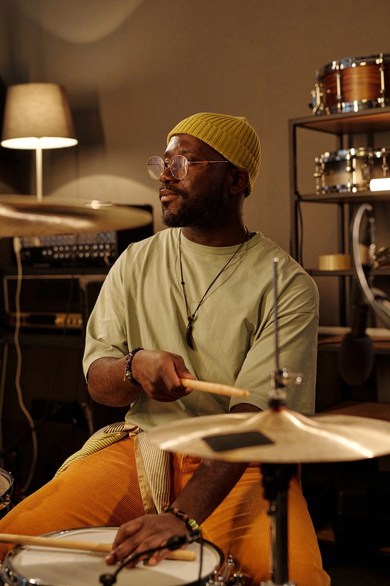 Musician wearing yellow hat and eyeglasses, playing drums in indoor studio with cozy lighting, surrounded by music equipment and instruments, creating harmonious atmosphere