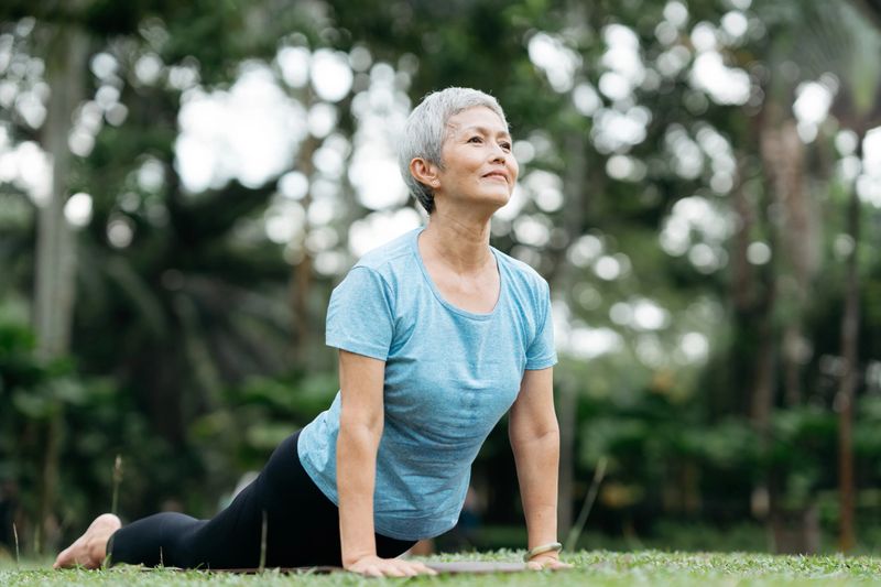 Senior Asian woman is practicing yoga in a park, doing stretching and relaxing exercises outdoors.
