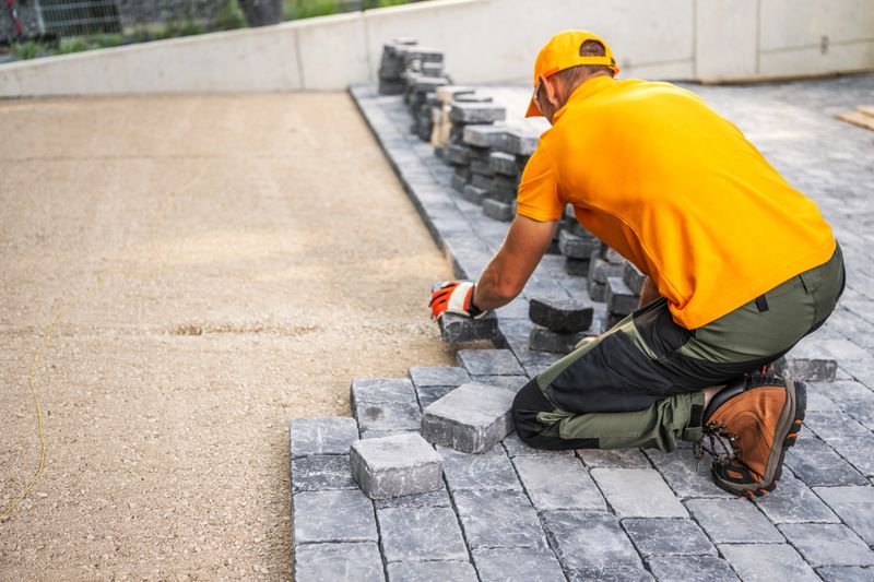 A construction worker carefully positions cobblestones to create a new patio in a residential yard under bright sunlight.