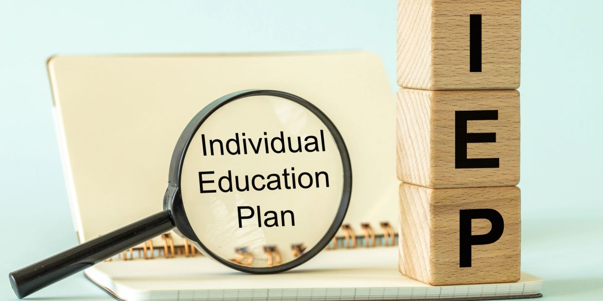 Magnifying glass highlighting 'Individual Education Plan' with wooden blocks spelling 'IEP'.