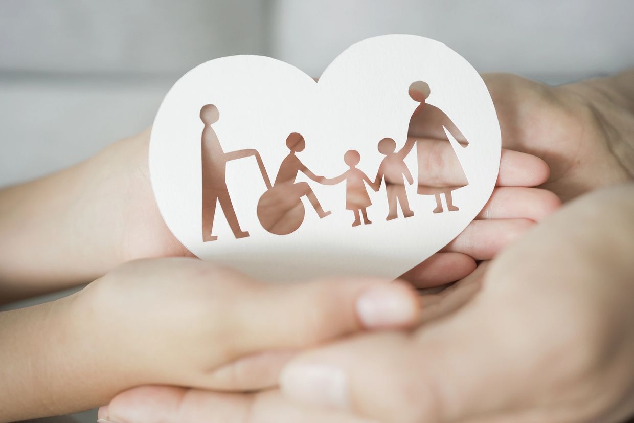 Hands holding a heart-shaped paper cutout of a diverse family with disability.