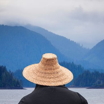 Person in a woven hat gazing at misty blue mountains across water.