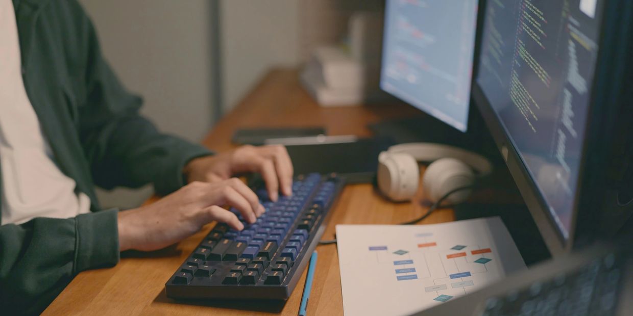 Person typing code on a mechanical keyboard at a desk with dual monitors and headphones.