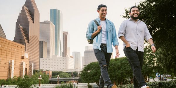 Two men walking and talking in a city park with skyscrapers in the background.