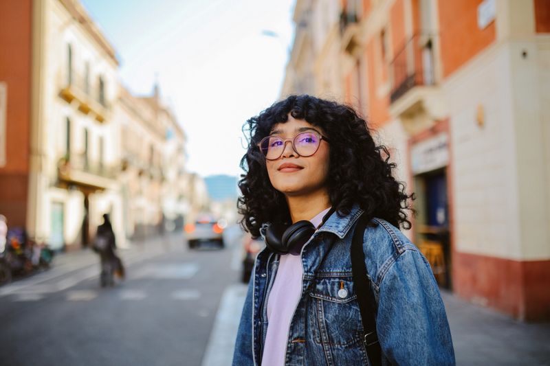 Fashionable young woman with curly hair and glasses stands on a street in Barcelona, wearing headphones and a denim jacket, enjoying the day.