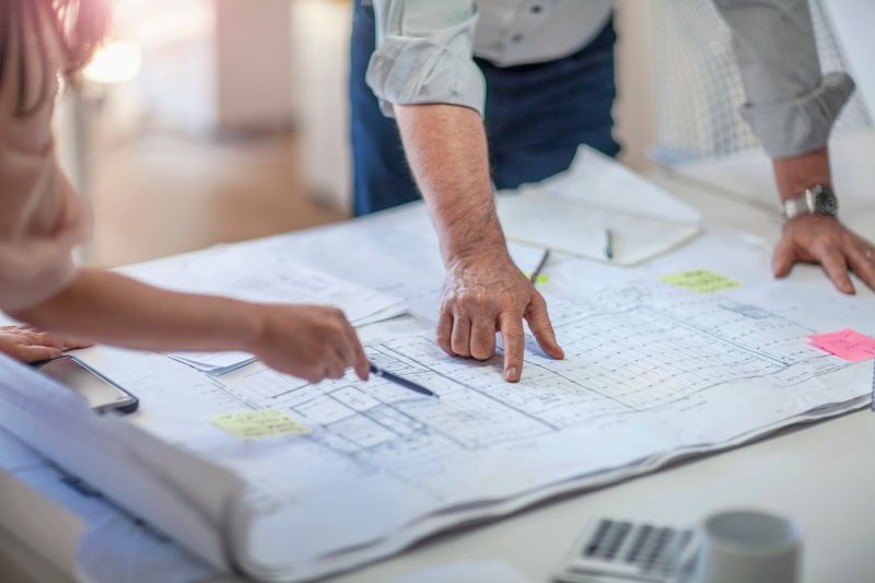 Colleagues gesturing pointing at architectural plans on tabletop desk in office