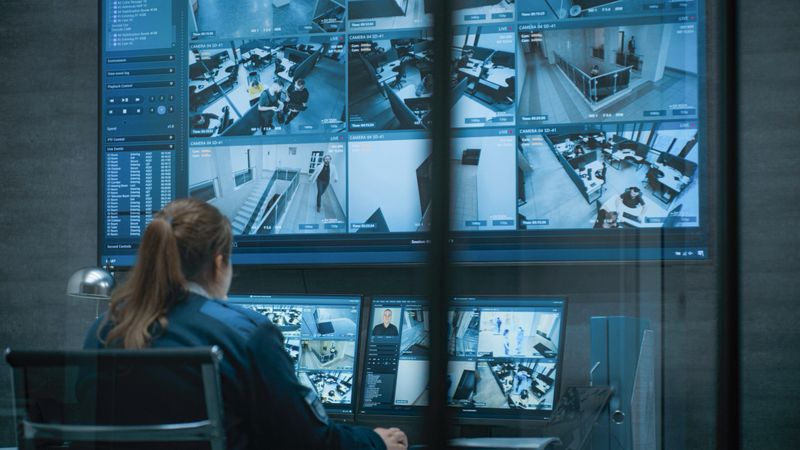 Female CCTV operator works on computer and tablet in monitoring center with security cameras, talks on walkie talkie. Surveillance video footage displayed on multiple monitors and big digital screen.
