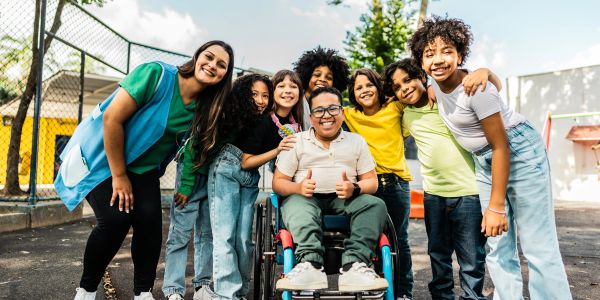 Group of happy children and a teacher outdoors, with one child in a wheelchair.