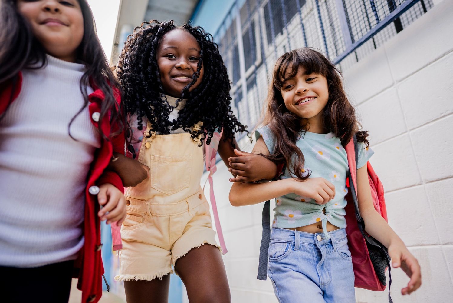 Three young girls walking together outdoors, smiling and holding arms.