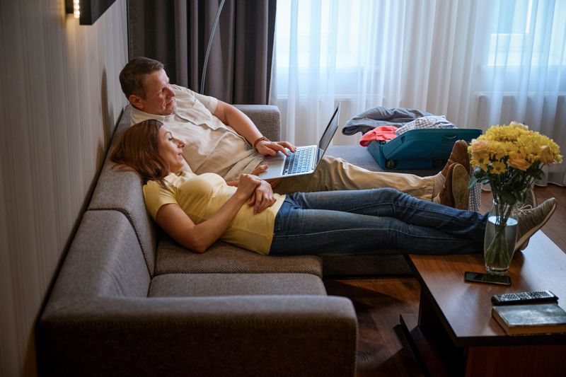 Hotel guests spend time together on a cozy sofa with a laptop, an open travel suitcase nearby