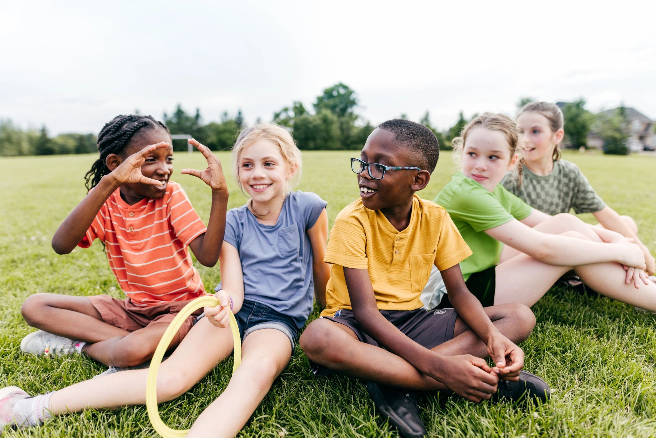 Five children sitting on grass, chatting and smiling during outdoor playtime.